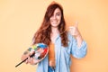 Young beautiful woman holding paintbrush and palette smiling happy and positive, thumb up doing excellent and approval sign Royalty Free Stock Photo