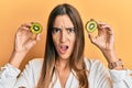 Young beautiful woman holding slice of kiwi over eyes in shock face, looking skeptical and sarcastic, surprised with open mouth Royalty Free Stock Photo