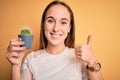 Young beautiful woman holding small cactus plant pot over isolated yellow background happy with big smile doing ok sign, thumb up Royalty Free Stock Photo