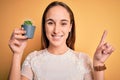 Young beautiful woman holding small cactus plant pot over isolated yellow background surprised with an idea or question pointing Royalty Free Stock Photo