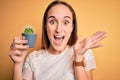 Young beautiful woman holding small cactus plant pot over isolated yellow background very happy and excited, winner expression Royalty Free Stock Photo