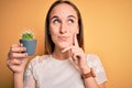 Young beautiful woman holding small cactus plant pot over  yellow background serious face thinking about question, very Royalty Free Stock Photo