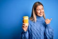 Young beautiful woman holding take away glass of coffee over isolated blue background pointing and showing with thumb up to the Royalty Free Stock Photo