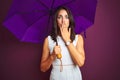 Young beautiful woman holding umbrella standing over purple isolated background cover mouth with hand shocked with shame for Royalty Free Stock Photo
