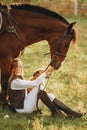 A young beautiful woman jockey with her dog sits in a meadow near her horse at sunset. Royalty Free Stock Photo