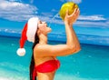 Young beautiful woman with long black hair in red bikini, dressed in red Santa Claus hat is drinking coconut water on tropical be