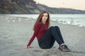 Young beautiful woman with long hair, in black jeans and red shirt, sitting on sand on beach among seagulls birds Royalty Free Stock Photo