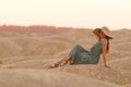 Young beautiful woman with long hair woith straw hat and in dress sitting elegant on sand at sunrise in sandy desert Royalty Free Stock Photo