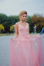 Young beautiful woman in long pink evening dress walking path in park. Fashion style portrait of gorgeous beautiful girl outdoors Royalty Free Stock Photo