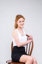 Young beautiful woman with long red, curly hair sitting on a wooden chair on a white background in the studio. Dressed in a white Royalty Free Stock Photo