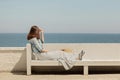 Young beautiful woman in a long skirt sits on a bench on a background of the sea horizon.