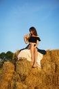 Young beautiful woman looks at the setting sun and holds a straw hat, sits on a large pile of straw bales Royalty Free Stock Photo