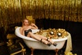 A young beautiful woman lying in a bathtub against the background of a golden interior holds a martini glass in a Royalty Free Stock Photo