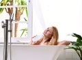 Woman lying in bathtub taking bath near open bathroom window and wash hand with soft shower gel cream lotion Royalty Free Stock Photo