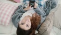 Young beautiful woman lying on the bed and watches photos on smartphone. Girl looks to the camera and smile.