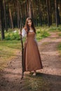 Young beautiful woma in medieval cowboy clothes, with a stick in hand. Barefoot on the ground. Against the background of the fores