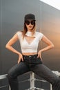 Young beautiful woman model in fashion sunglasses in sexy trendy torn white t-shirt in jeans and cool baseball cap sit on metal Royalty Free Stock Photo