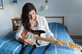 Young beautiful woman, musician, singer studying, practicing to play electric guitar at home sitting on bed in blue shirt,jeans. Royalty Free Stock Photo