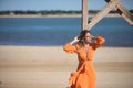 Young and beautiful woman in an orange dress, leaning on a wooden pole, hands on her head, looking at infinity, relaxed and calm, Royalty Free Stock Photo