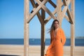 Young and beautiful woman in an orange dress, leaning on a wooden post, relaxed and calm, looking at the camera, in solitude.