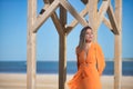 Young and beautiful woman in an orange dress, leaning on a wooden post, relaxed and calm, looking at infinity, in solitude.