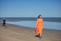 Young and beautiful woman in an orange dress and pink heels in her hand, walking barefoot on the beach in solitude. Concept beauty