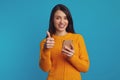 Young beautiful woman in orange sweater showing thumb up while using smartphone Royalty Free Stock Photo