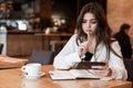 Young beautiful woman with pen in her hand taking notes to her planner using her smartphone looks hesitant drinking hot coffee in