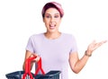 Young beautiful woman with pink hair holding supermarket shopping basket celebrating victory with happy smile and winner