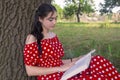 Young beautiful woman in red dress reads a book in the park. The lady with her back to the tree trunk in the summer outdoors Royalty Free Stock Photo