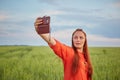 Young beautiful woman in red dress and red hair, takes a selfie on the phone in the green wheat field in the evening at Royalty Free Stock Photo