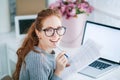 Young beautiful woman with red hair, wearing glasses, working in the office, uses a laptop and mobile phone Royalty Free Stock Photo