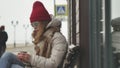 Young beautiful woman in a red hat wearing sporty warm clothes and rollers, sitting on a wooden bench drinking tea from Royalty Free Stock Photo