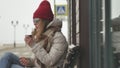 Young beautiful woman in a red hat wearing sporty warm clothes and rollers, sitting on a wooden bench drinking tea from Royalty Free Stock Photo