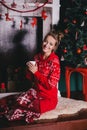 Young beautiful woman in a red warm pajamas with scandinavian ornaments sitting near decorative fireplace and drinking hot tea Royalty Free Stock Photo