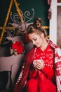 Young beautiful woman in a red warm pajamas with scandinavian ornaments sitting near decorative fireplace and drinking hot tea Royalty Free Stock Photo