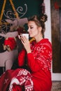 Young beautiful woman in a red warm pajamas with scandinavian ornaments sitting near decorative fireplace and drinking hot tea Royalty Free Stock Photo