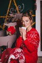 Young beautiful woman in a red warm pajamas with scandinavian ornaments sitting near decorative fireplace and drinking hot tea Royalty Free Stock Photo