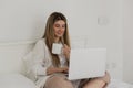 Young beautiful woman relaxing and drinking cup of hot coffee or tea using laptop computer on the bed in the bedroom. Royalty Free Stock Photo