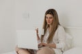 Young beautiful woman relaxing and drinking cup of hot coffee or tea using laptop computer on the bed in the bedroom. Royalty Free Stock Photo