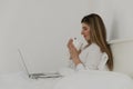 Young beautiful woman relaxing and drinking cup of hot coffee or tea using laptop computer on the bed in the bedroom. Royalty Free Stock Photo