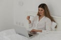 Young beautiful woman relaxing and drinking cup of hot coffee or tea using laptop computer on the bed in the bedroom. Royalty Free Stock Photo