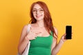 Young beautiful woman showing to camera blank screen of smart phone isolated over yellow background, being in good mood, red Royalty Free Stock Photo