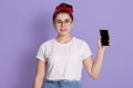 Young beautiful woman shows right in camera blank screen of smart phone isolated over lilac background, girl in white casual t Royalty Free Stock Photo