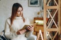 A young beautiful woman sits in a chair, covered with a blanket, reading a book near the window on Christmas Eve. Royalty Free Stock Photo