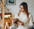 A young beautiful woman sits in a chair, covered with a blanket, reading a book near the window on Christmas Eve. Royalty Free Stock Photo