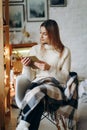 A young beautiful woman sits in a chair, covered with a blanket, reading a book near the window on Christmas Eve. Royalty Free Stock Photo