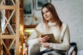 A young beautiful woman sits in a chair, covered with a blanket, reading a book near the window on Christmas Eve. Royalty Free Stock Photo