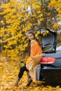 A young beautiful woman sits on the trunk of a car in the autumn Sunny forest and smiles. The concept of autumn travelling