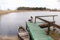 Young beautiful woman sits on the wooden bridge on the river at spring day. old boat near the shore. selective focus Royalty Free Stock Photo